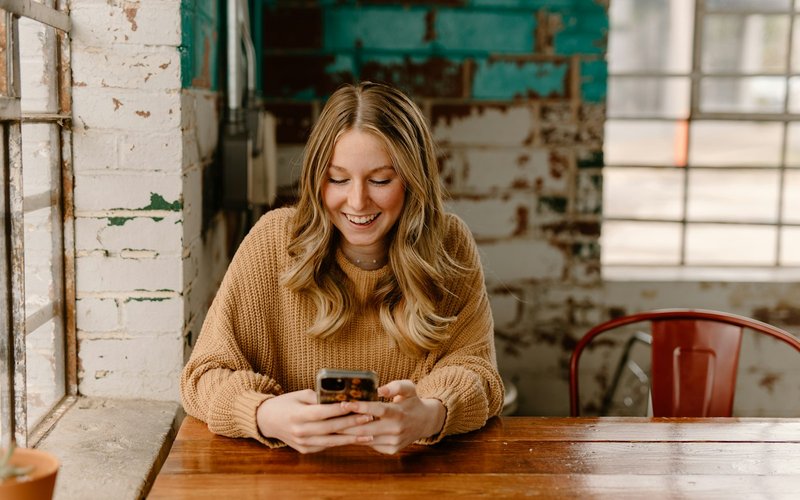Une femme souriante consulte son téléphone attablée dans un café.
