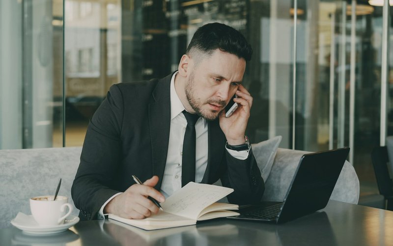 Un homme en costume parle au téléphone tout en prenant des notes dans un carnet.
